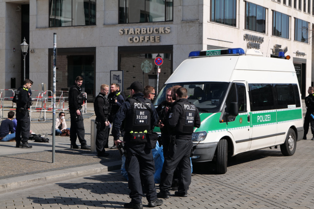 Polizisten vor einem Starbucks-Café mit einem Van auf der rechten Seite und Menschen auf der linken Seite vor einem Hintergrund mit Fenstern, einer Tafel, einem Laternenmast und einem Zaun.