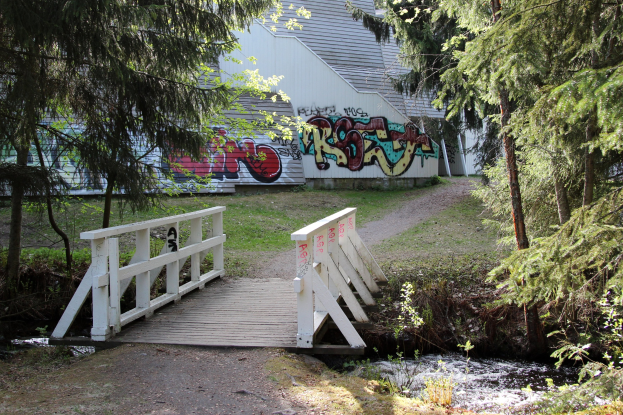 Holzbrücke über einen Bach vor einem Graffiti-bedeckten Gebäude, umgeben von grünem Gras und Bäumen.