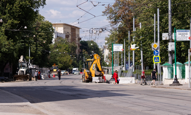 Eine Stadtstraße mit einer Baustelle, Fahrzeugen, Fußgängern, einem Radfahrer, Verkehrsleitkegeln, Pfosten, Hinweisschildern, Strommasten mit Kabeln, Bäumen, Gebäuden mit Fenstern und einem bewölkten Himmel.