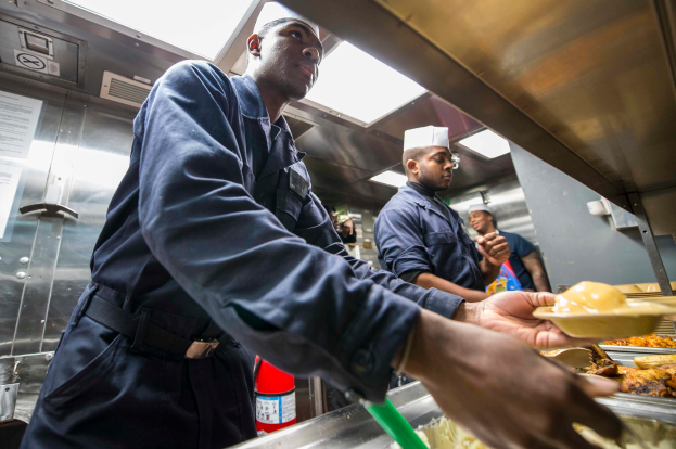 Eine Gruppe Von M├Ąnnern Steht In Einer K├╝che, Einer Hält Ein Tablett Mit Essen, Mit Tabletts Von Essen Auf Dem Tisch, Einem Feuerl├Âscher Und Papieren An Der Wand Im Hintergrund.