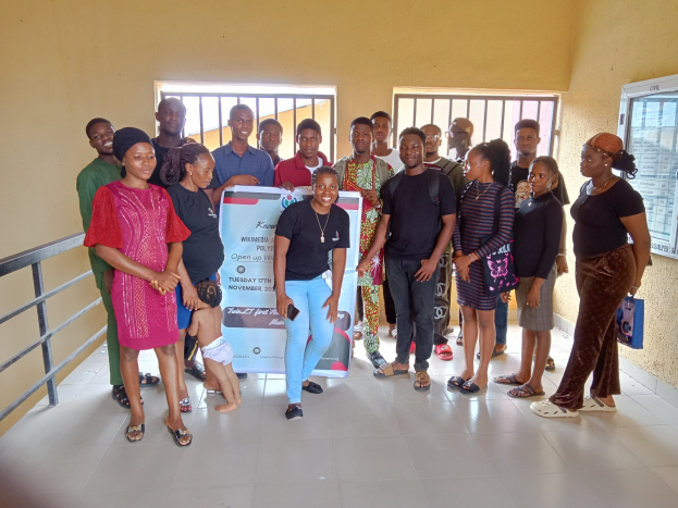 Eine Gruppe von Menschen, die zusammen in einem Raum stehen und eine Informationswand halten, mit einem Geländer auf der linken Seite, Fenstern und einer Wand im Hintergrund.