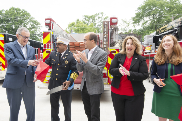 A group of people clapping and smiling during a ribbon cutting ceremony in front of a fire truck, with two individuals holding scissors and a red ribbon.