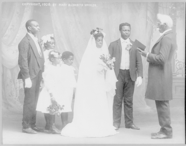 Ein Schwarz-Weiß-Foto einer Braut in einem weißen Kleid und Schleier, die einen Strauß hält und neben einem Bräutigam steht, der ein Buch hält, mit ihrer Familie und Vorhängen im Hintergrund.