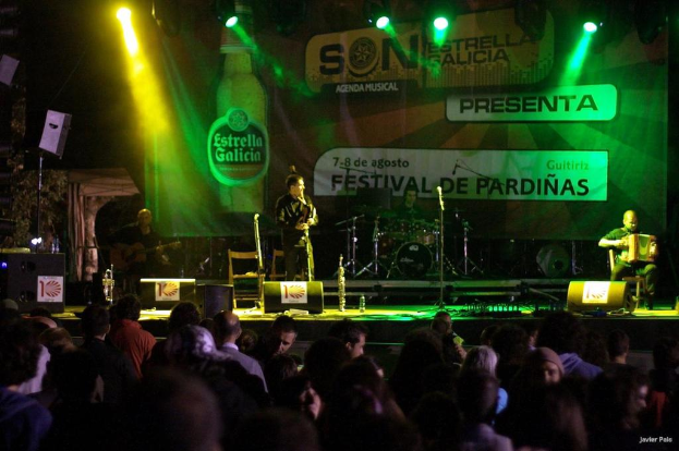 Ein Publikum schaut einer Vorstellung auf der Bühne zu, auf der Musiker Instrumente spielen, Lautsprecher, Mikrofone, Werbetafeln und fokussiertes Licht zu sehen sind, sowie eine Flasche und andere Gegenstände im Hintergrund.