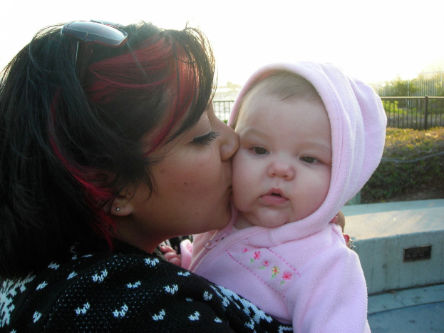 Eine Frau umarmt ein Baby in der Mitte des Bildes, mit Büschen und Zaun im Hintergrund und Boden in der rechten unteren Ecke sichtbar.