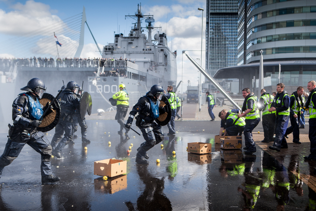 Eine Gruppe von Polizisten in Einsatzausrüstung, Helmen und mit Gegenständen in der Hand geht eine Straße entlang neben einem großen Schiff. Im Hintergrund sind verstreute Kartons, Gebäude, Pfosten, Fahrzeuge, Menschen, eine Brücke, eine Flagge und ein bewölkter Himmel zu sehen.