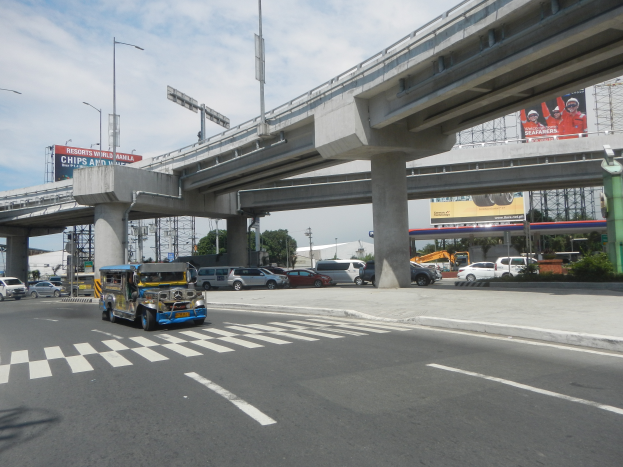 Eine vielbefahrene Straße mit Fahrzeugen, einer Zebrastreifen, einer Überführung mit Säulen, Laternen, Schildern mit Text und Bildern, Bäumen, Gebäuden und einem bewölkten Himmel im Hintergrund.