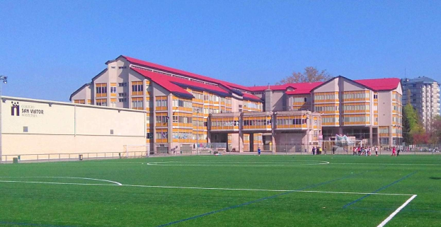 Ein Fußballfeld mit einem großen Gebäude im Hintergrund, das als San Visitor Academy beschriftet ist, umgeben von Bäumen und Laternen, mit ein paar Menschen auf dem Feld und dem Himmel darüber.