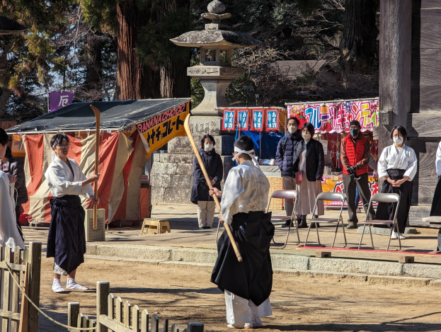 Eine Gruppe von Menschen in formeller Kleidung, einige mit Masken und Holzstöcken, versammeln sich im Freien vor einem Gebäude während einer traditionellen japanischen Zeremonie in Kyoto, mit Stühlen, einem Zelt, Bannern, Bäumen und einem klaren blauen Himmel im Hintergrund.