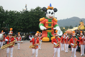 Eine Gruppe von Menschen in Kostümen spielt Musikinstrumente in der Nähe eines Mickey-Maus-Floats in einem Umzug, mit Bäumen, Gebäuden und einem klaren Himmel im Hintergrund und einer Tafel mit Text auf der linken Seite.
