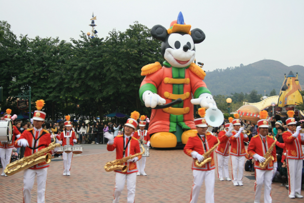 Eine Gruppe von Menschen in Kostümen spielt Musikinstrumente in der Nähe eines Mickey-Maus-Floats in einem Umzug, mit Bäumen, Gebäuden und einem klaren Himmel im Hintergrund und einer Tafel mit Text auf der linken Seite.