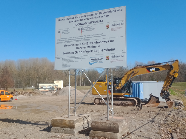 Eine Baustelle mit einem Bagger und einem Schild mit der Aufschrift "Rheinland-Pfalz - Hochwasserschutz", ein Fahrzeug auf der linken Seite und Bäume mit einem klaren blauen Himmel im Hintergrund.