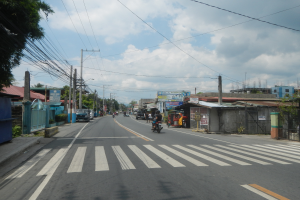 Eine Stadtstraße mit Fahrzeugen, einer Zebrastreifen, Strommasten mit Drähten, Gebäuden, Bäumen und einem bewölkten Himmel.