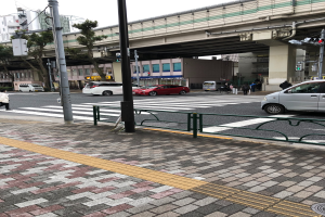 Eine Straßenszene in der Stadt mit Fahrzeugen, Fußgängern an einer Ampel, einer Brücke, Bäumen, Gebäuden und einem klaren blauen Himmel.