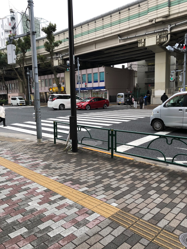 Eine Straßenszene in der Stadt mit Fahrzeugen, Fußgängern an einer Ampel, einer Brücke, Bäumen, Gebäuden und einem klaren blauen Himmel.