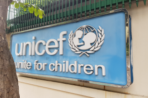 Das UNICEF-Logo ist auf einer Gebäudewand zu sehen, mit einem Baumstamm links daneben und einer an der Wand befestigten Tafel, vor einem Hintergrund aus Bäumen und einem klaren blauen Himmel.