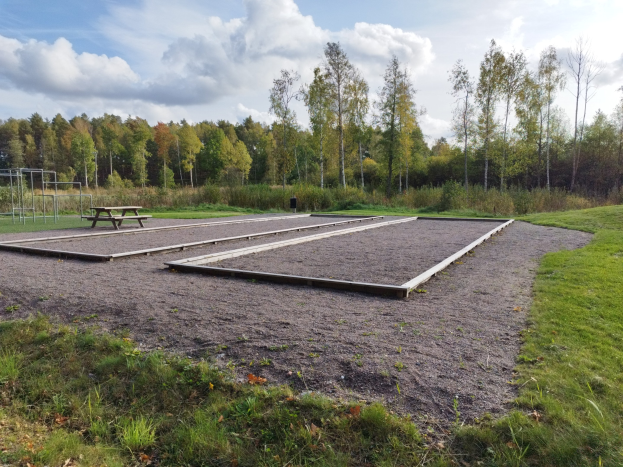 Eine grüne Wiese mit einem Picknicktisch, umgeben von Bäumen unter einem klaren blauen Himmel, mit Spielplatzgeräten einschließlich Stangen und einer Bank auf der linken Seite.