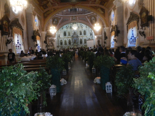Eine Hochzeitszeremonie in einer Kirche mit Gästen auf Bänken sitzend und im Gang stehend, dekoriert mit Zimmerpflanzen, Skulpturen, Vorhängen, Kronleuchtern und einem Bildschirm im Hintergrund.