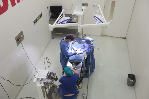 Chirurgen führen einen Eingriff in einem Operationssaal durch, umgeben von medizinischer Ausrüstung, einer Wand mit Text auf der linken Seite und einem Objekt auf dem Boden auf der rechten Seite.