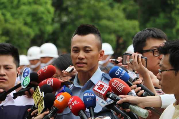 Ein Mann in einer Polizeiuniform steht umgeben von Reportern mit Mikrofonen und Mobiltelefonen, mit Bäumen im Hintergrund.