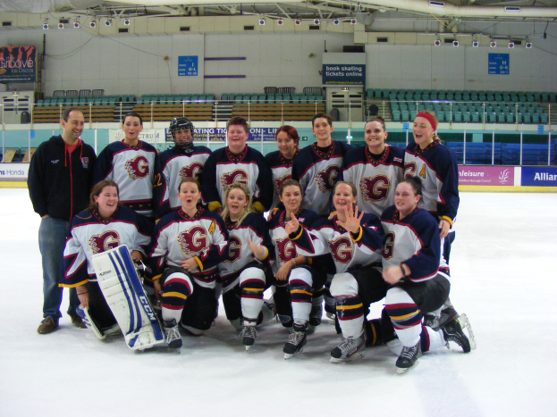 Eine Gruppe von Frauen-Eishockeyspielerinnen in Helmen und Schlittschuhen posiert für ein Foto auf dem Eis, eine hält einen Hockey-Schläger, mit Stühlen, Tafeln mit Text, Geländern und Deckenlampen im Hintergrund.