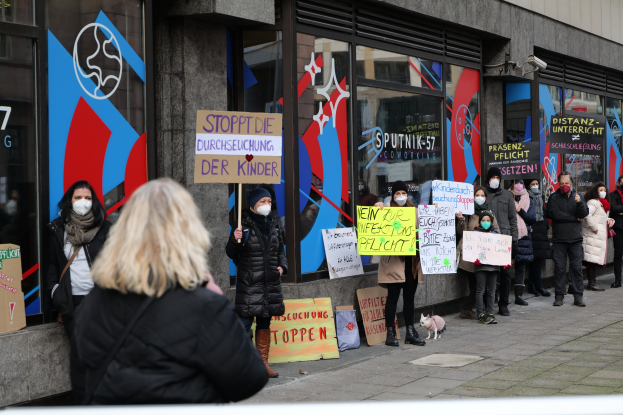 Eine Gruppe maskierter Menschen mit Schildern und Protestschildern vor einem Glasfrontgebäude, mit Kameras und einem Hund unten.