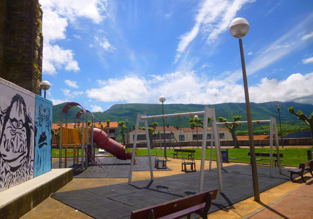 Ein Spielplatz mit Schaukeln, einer Rutsche, Bänken, Masten, Lichtern, einer bemalten Wand, Gras, Bäumen, Gebäuden, Hühlen und einem bewölktem Himmel im Hintergrund.