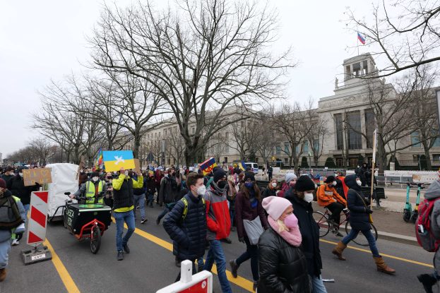 Eine großle Gruppe von Menschen marschiert bei einer Demonstration durch eine Straße in Washington, D.C., mit Schildern und Bannern, während einige Fahrräder fahren, unter einem klaren blauen Himmel.