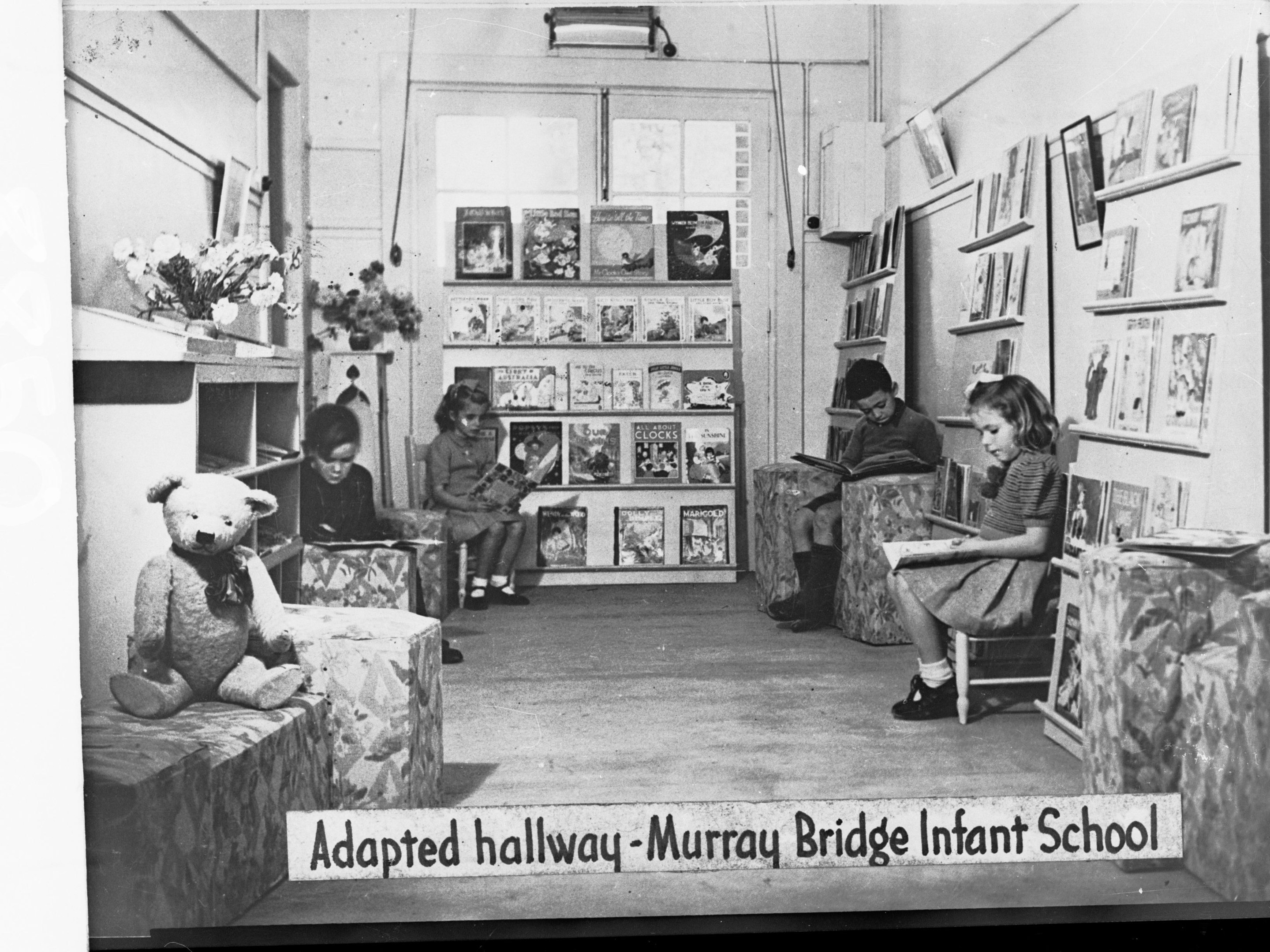 Eine Gruppe von Kindern sitzt auf Stühlen in einem Raum mit Bücherregalen, einem Teddybär links, einer Vase auf dem Tisch und gerahmten Gegenständen an der Wand, mit dem Text "Angepasster Flur Murray Bridge Kindergarten" unten.