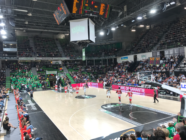 Ein Basketballspiel in einer großen Arena, mit Zuschauern auf den Rängen und Anzeigetafeln, die "Asvel vs Galatasaray" anzeigen.