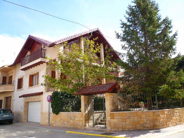 Eine Villa mit einem davor geparkten Auto, mit Fenstern, Geländern, Pfeilern und einem Dach, umgeben von einem Tor, einem Schild, einem Straßenschild, Bäumen und einer Wand unter einem bewölkten Himmel.