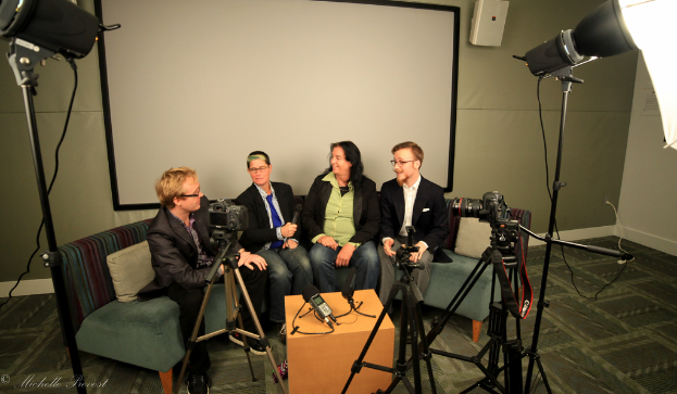 Eine Gruppe von drei Personen sitzt auf einem Sofa und gibt ein Interview, wobei eine Person links in einem schwarzen Mantel und Hose, eine Person in einem Mantel und blauer Krawatte mit einem Mikrofon in der Mitte und eine Person rechts lächelt. Ein Mikrofonrecorder befindet sich auf dem Mitteltisch, Kameras sind auf der rechten und linken Seite und große Scheinwerfer sind vorhanden, mit einem Projektionsschirm im Hintergrund.