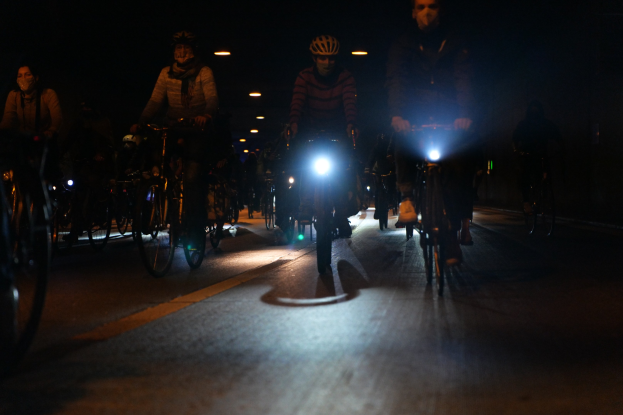 Eine Gruppe von Menschen, die nachts auf Fahrrädern eine gut beleuchtete Stadtstraße hinunterfahren, alle tragen Helme.