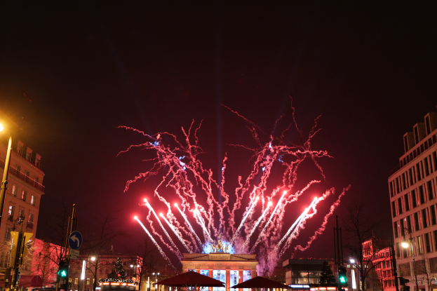Eine belebte StraßenSzene in Berlin an Silvester, voller Menschen, Fahrzeuge und Gebäude, beleuchtet von Feuerwerk und Lichtern, die eine festliche Atmosphäre schaffen.