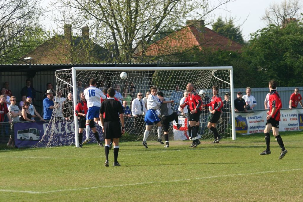 Fußballspieler spielen auf einem Feld mit einem Tor im Hintergrund, während Zuschauer zuschauen; im Hintergrund sind Bäume und Häuser zu sehen.