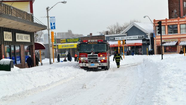 Ein Feuerwehrauto steht im Schnee vor Gebäuden, mit Geschäften und Laternen auf der linken Seite, einer Ampel in der Mitte und Schnee auf dem Boden.