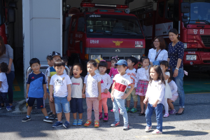 Eine Gruppe von Kindern vor einem Feuerwehrauto auf einer Feuerwache, einige tragen Mützen, mit zusätzlichen Feuerwehrautos im Hintergrund.