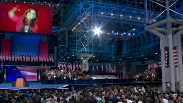 Eine große Menschenmenge sitzt vor einer Bühne mit einem Podium, einem Mikrofon, Flaggen, einem Bildschirm mit dem Bild einer Frau und Lichtausrüstung, wahrscheinlich bei einer Präsidentschaftsdiskussion.