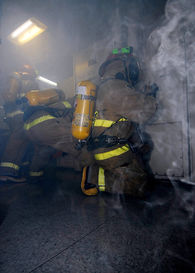 Feuerwehrleute in Schutzausrüstung mit Rauch aus einer Tür, beleuchtet von einer Lichtquelle an einer Wand.