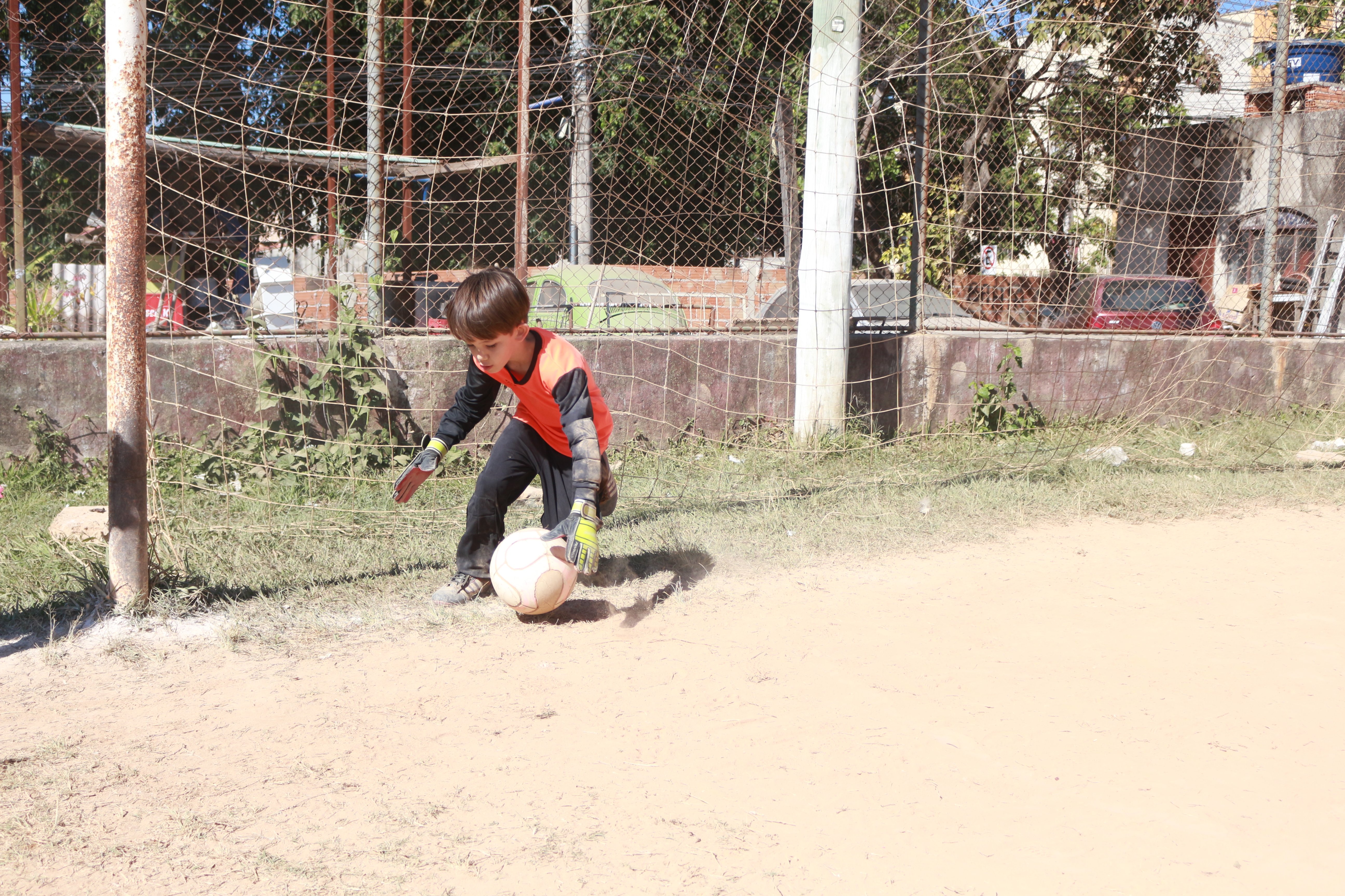 Ein junger Junge spielt Fußball auf einem Schotterfeld, trägt Schuhe und hat verschiedene Elemente wie Gras, Pflanzen, Pfosten, einen Zaun, eine Wand, Bäume, Fahrzeuge, Gebäude und einen bewölkten Himmel im Hintergrund.