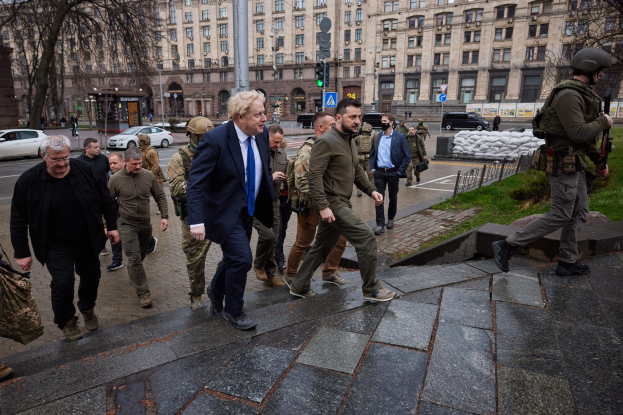 Boris Johnson erreicht das Kreml in Moskau, geht eine Straße entlang an einem Gebäude vorbei, während andere Helme tragen und Waffen halten, während Fahrzeuge, Laternenmasten, Verkehrsampeln, Bäume und Gebäude mit Fenstern den Hintergrund bilden.
