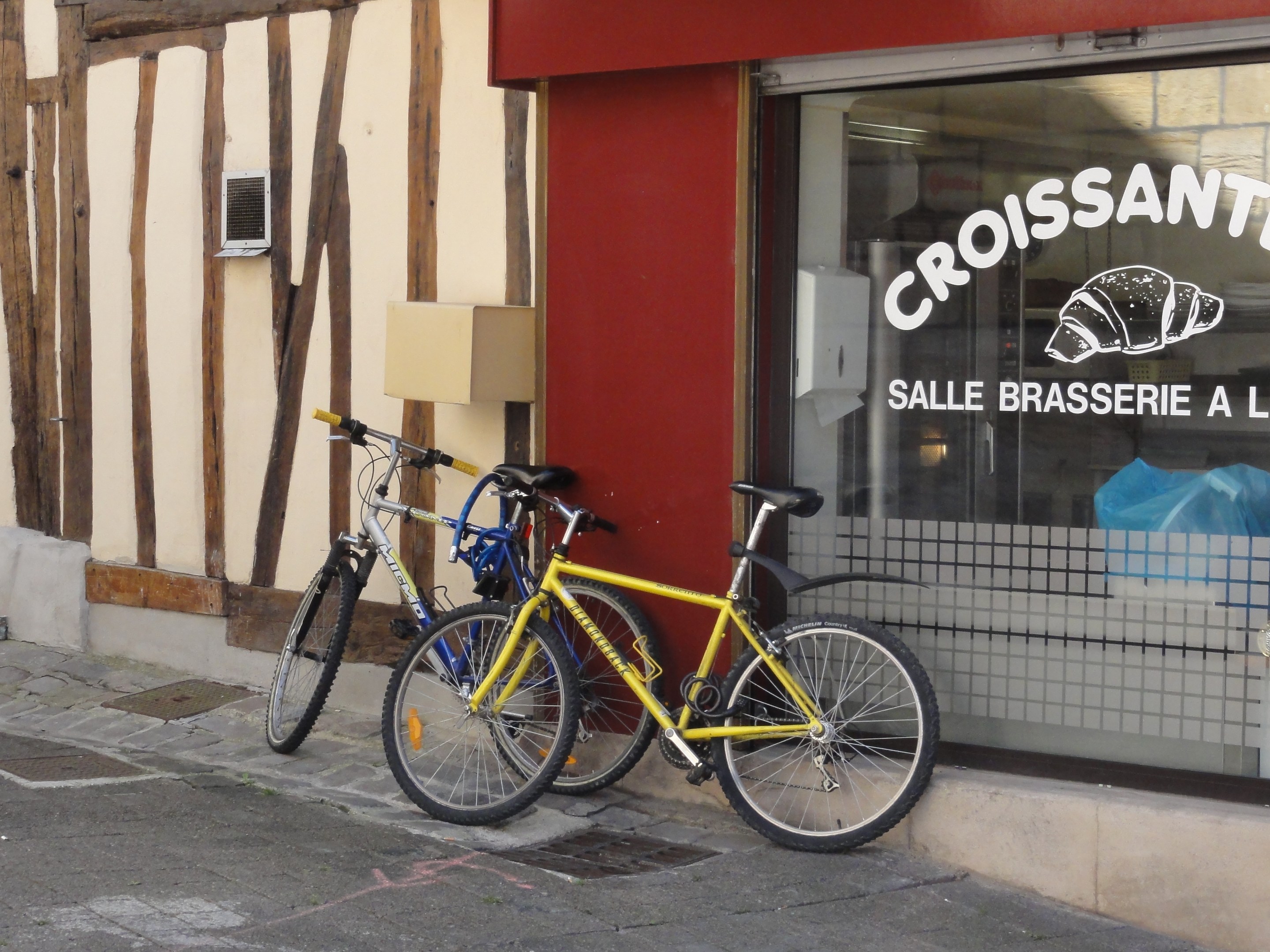 Zwei Fahrräder nebeneinander auf einem Gehweg vor einem Geschäft mit einer Glaswand mit Text und einem Objekt links von den Fahrrädern.