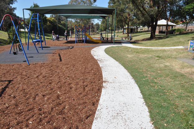 Ein Park mit einem Spielplatz, der Schaukeln und Rutschen bietet, umgeben von grünem Gras, Bäumen, Bänken, Hütten und einem klaren blauen Himmel.