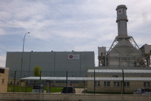 Ein großes Gebäude mit einem zentralen Turm, umgeben von einem Zaun, mit Fahrzeugen auf der benachbarten Straße, Masten, Lampen, Drähten, Bäumen und Gras im Vordergrund und einem bewölkten Himmel im Hintergrund, das eine neue Energiespeicheranlage darstellt.