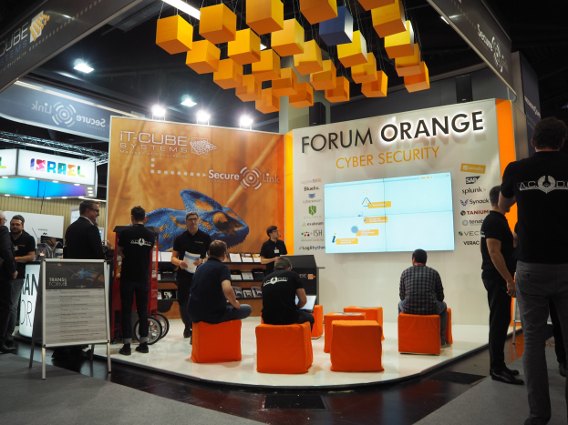 Eine Gruppe von Menschen an einem IT-Cube Systems Forum Orange Cyber Security Stand auf einer Messe, mit Stühlen, einer Tafel, einem Display-Bildschirm, Regalen, Schildern, Deckenleuchten und Lampen.