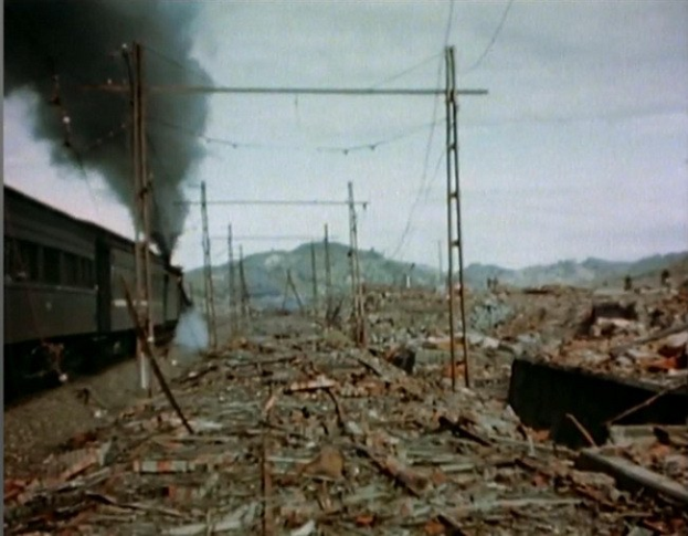 Ein Zug fährt durch ein beschädigtes Gebiet mit Rauch, der aus ihm aufsteigt, umgeben von Strommasten und -leitungen, mit Hügeln und einem klaren Himmel im Hintergrund und verstreuten Trümmern auf dem Boden.