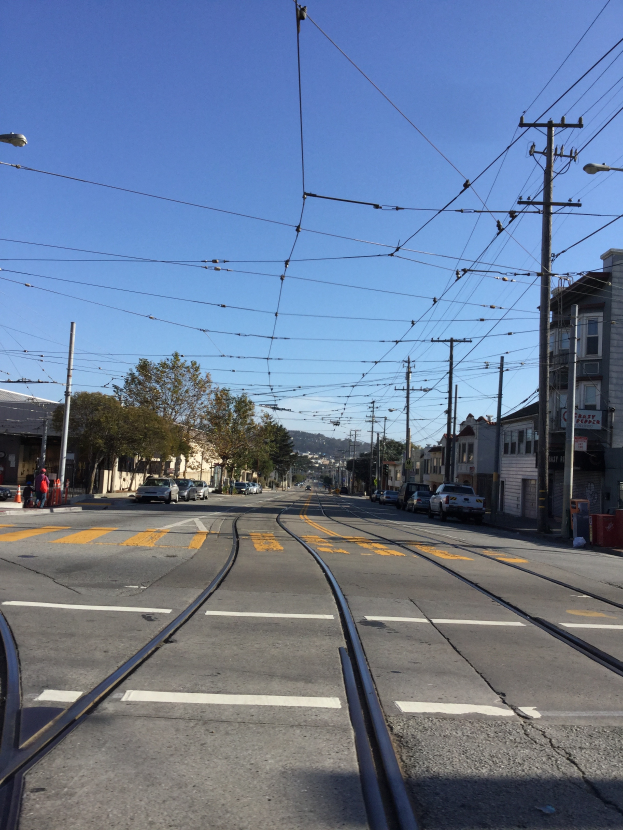Eine Stadtstraße mit einer Bahnschiene, Fahrzeugen, Strommasten, Laternen, Bäumen, Gebäuden mit Fenstern, einigen Menschen, Hügeln und einem klaren blauen Himmel im Hintergrund.