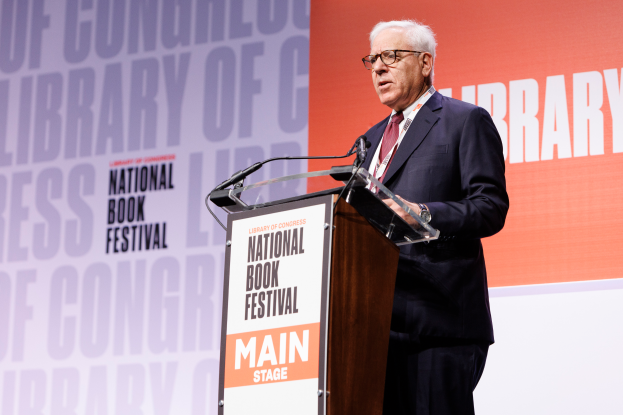 Ein Mann im Blazer, Krawatte und Brille spricht in ein Mikrofon an einem Podium vor einer 'National Book Festival'-Tafel.