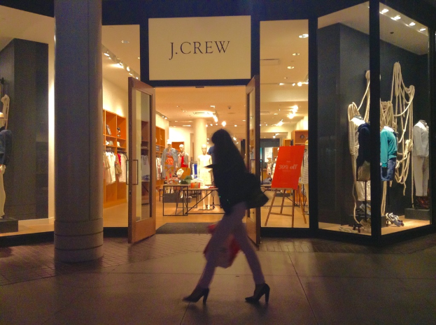 Ein Einkaufszentrum mit Glaswänden und -türen, das Puppen, Tische, hängende Kleider und Schränke sowie eine Frau zeigt, die daneben geht.