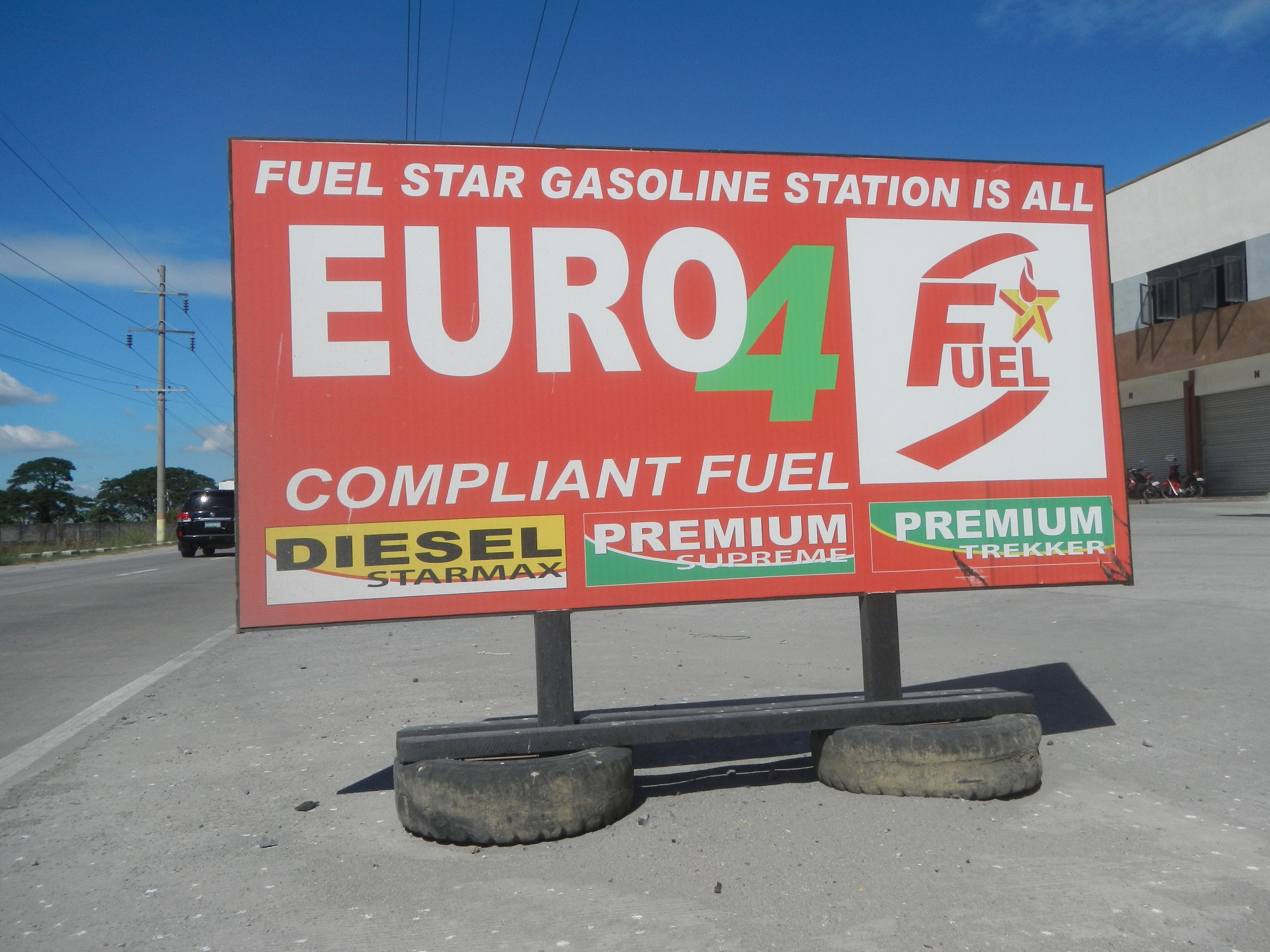 Tankstelle mit dem Schild "Euro4 Fuel", Fahrzeuge auf der Straße, Strommasten mit Drähten, Bäume, ein Gebäude auf der rechten Seite und sichtbarer Himmel.